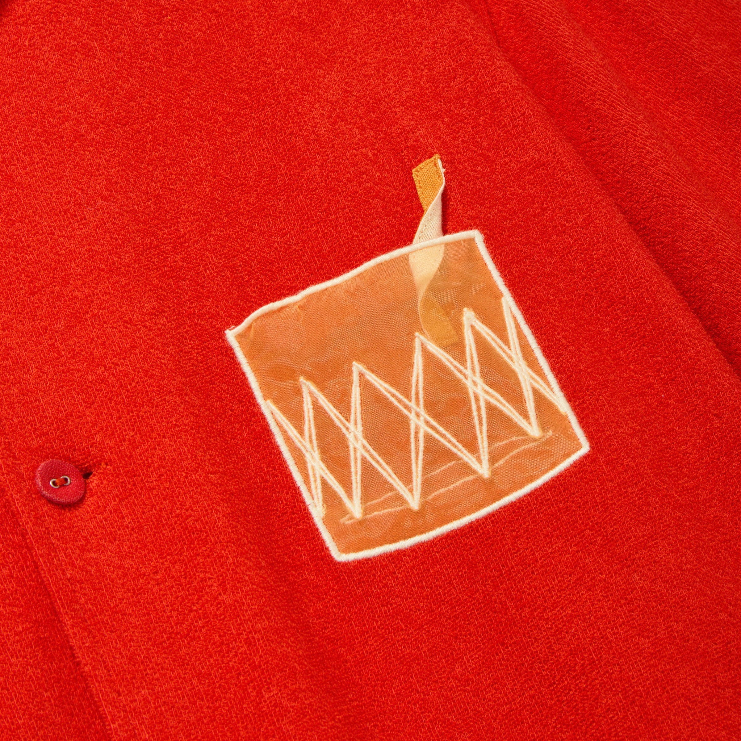 Close-up of a red cabana shirt showing an embroidered Negroni glass on the chest pocket, featuring a transparent mesh panel and a stitched orange garnish detail.