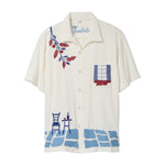 Cream terry cabana shirt with embroidered blue and red Mediterranean motifs, including flowers, a window, café table, chair, and cobblestone hem.