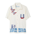 Cream terry cabana shirt with embroidered blue and red Mediterranean motifs, including flowers, a window, café table, chair, and cobblestone hem.