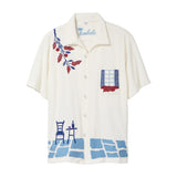 Cream terry cabana shirt with embroidered blue and red Mediterranean motifs, including flowers, a window, café table, chair, and cobblestone hem.