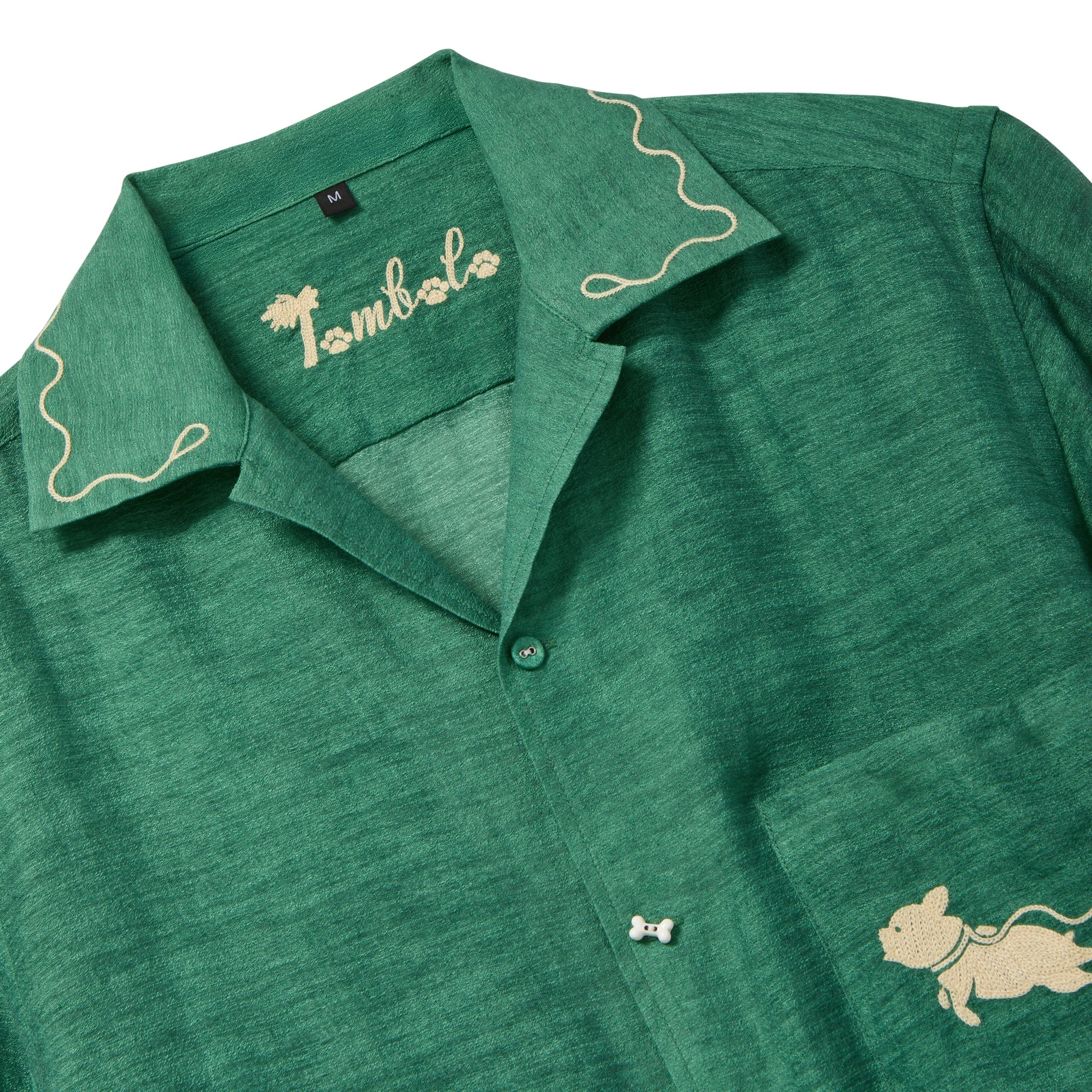 Close-up of green cabana shirt showing contrast wavy piping along the collar, Tombolo logo embroidered inside the neckline, chest pocket with embroidered dog graphic, and a dog bone button at the center front.