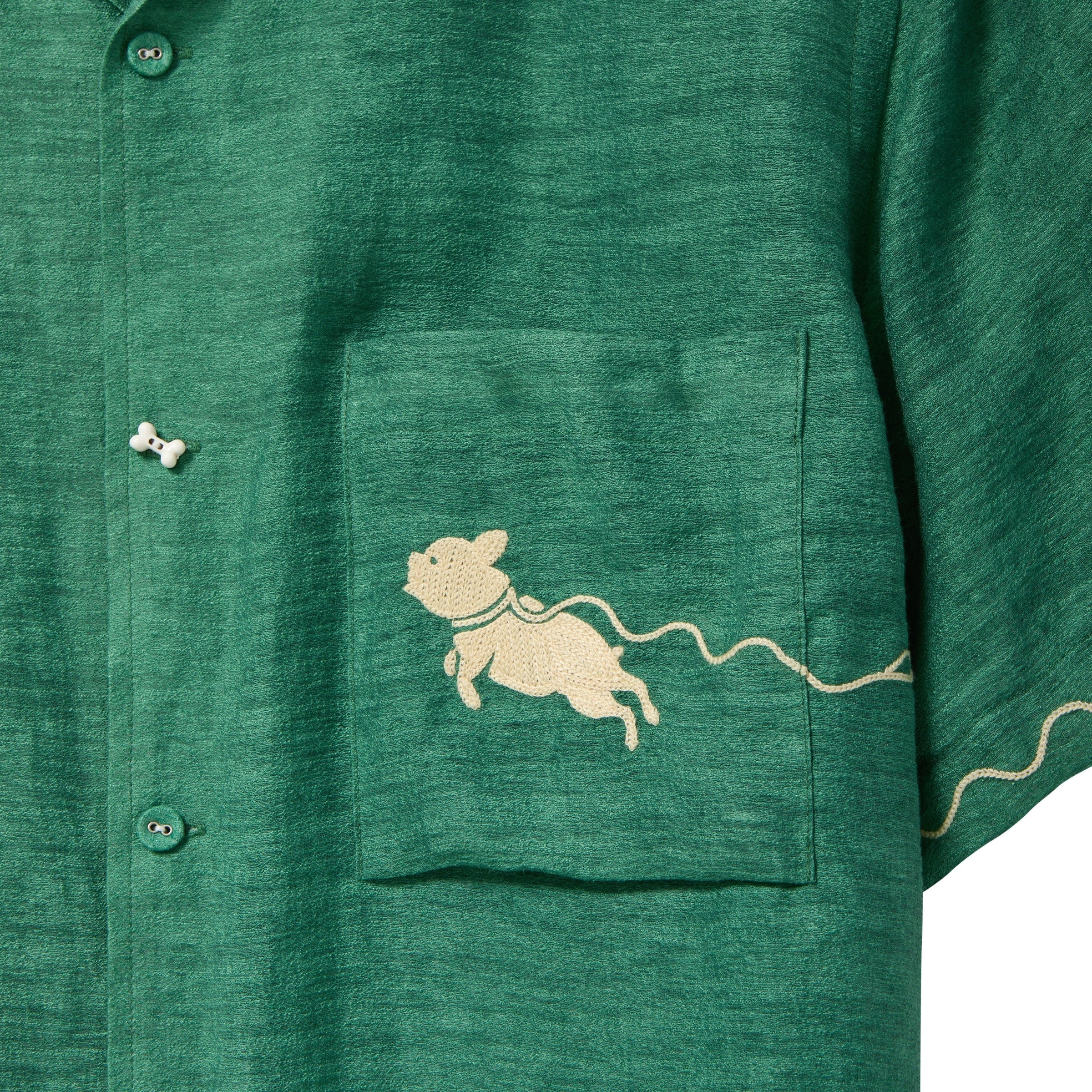Close-up of green cabana shirt showing chest pocket with embroidered dog graphic and dog bone button at the center front.