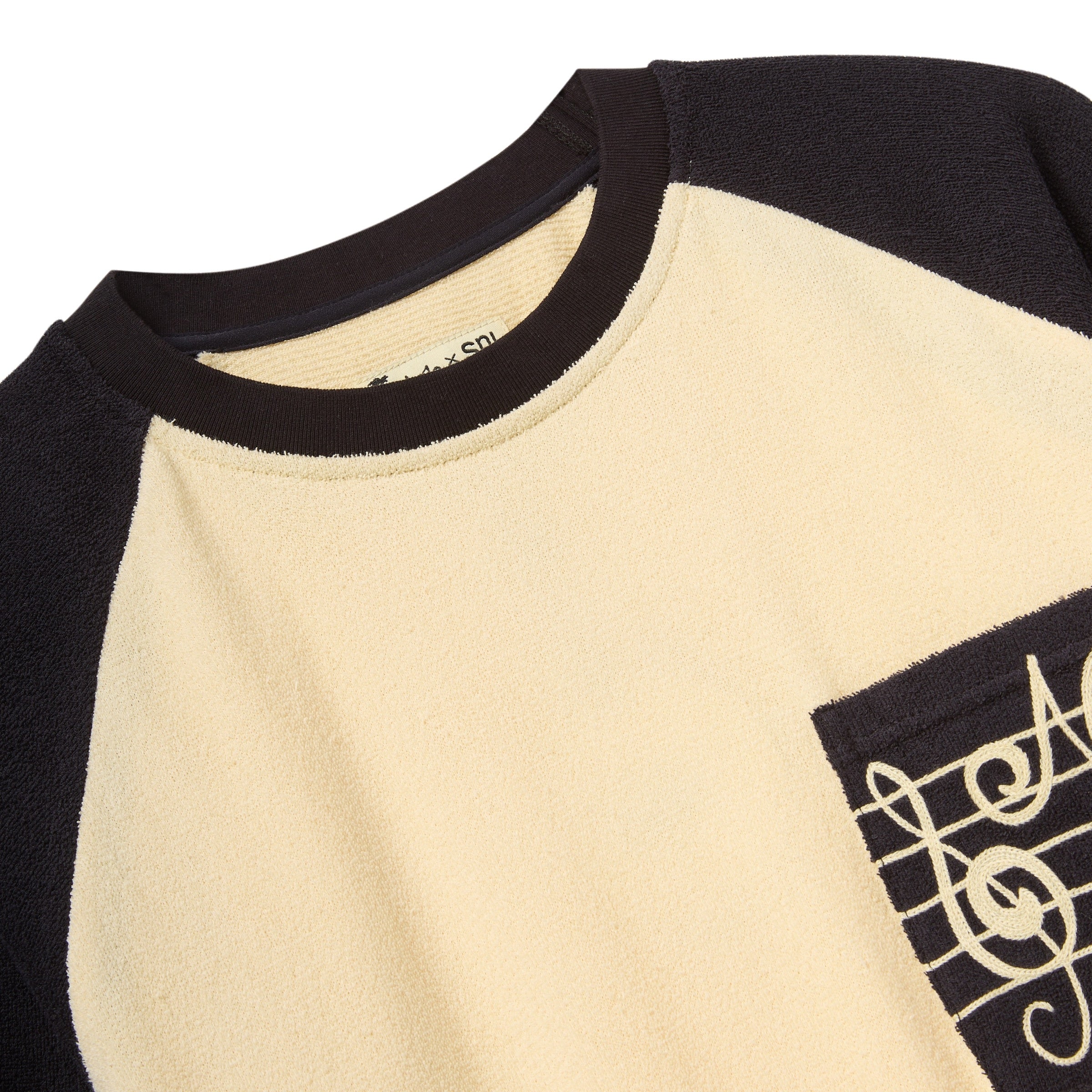 Close-up of a beige and black shirt with a music note SNL logo on the chest pocket. 