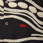 Close-up of black shirt with cream chain-stitch embroidery in dotted and wave patterns, featuring a small red stitched loon eye detail.