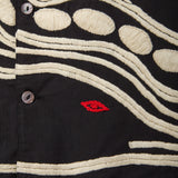 Close-up of black shirt with cream chain-stitch embroidery in dotted and wave patterns, featuring a small red stitched loon eye detail.