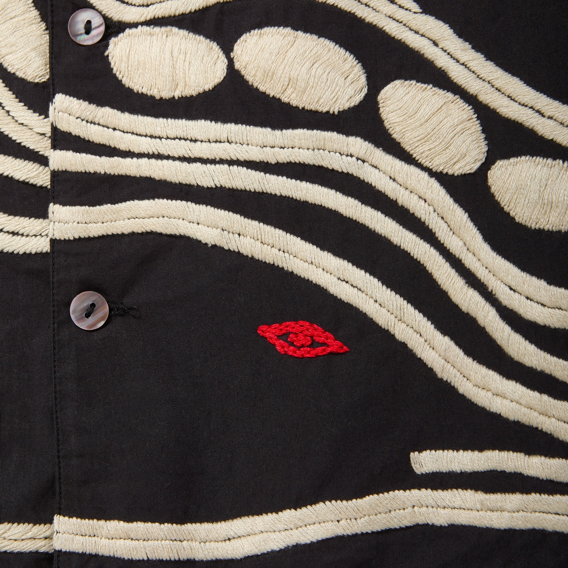 Close-up of black shirt with cream chain-stitch embroidery in dotted and wave patterns, featuring a small red stitched loon eye detail.