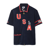 Navy terry cloth zip-front shirt with red contrast trim, oversized “USA” appliqué lettering, Statue of Liberty and FIFA World Cup patches, a chest pocket, sleeve graphics, and a gold-tone two-way zipper.