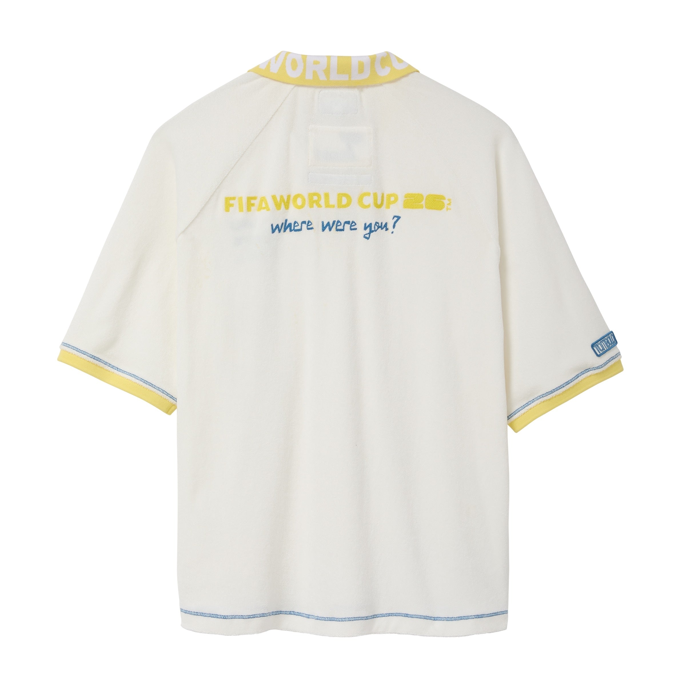Back of a cream terry cloth polo shirt with a yellow contrast collar, blue contrast stitching, yellow embroidered “FIFA World Cup 26™” text on the upper back, blue “where were you?” embroidery, and a FIFA World Cup sleeve patch.
