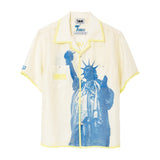 Cream camp collar shirt with yellow contrast trim, two chest pockets, a large blue Statue of Liberty graphic across the front, a FIFA World Cup 26 New York New Jersey logo on the pocket, and a FIFA World Cup sleeve patch.