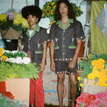 Man and woman standing in flower shop, each wearing brown picante shirt. The woman styles it with red pants. The man is wearing the matching shorts. 