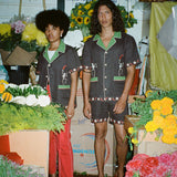 Man and woman standing in flower shop, each wearing brown picante shirt. The woman styles it with red pants. The man is wearing the matching shorts. 