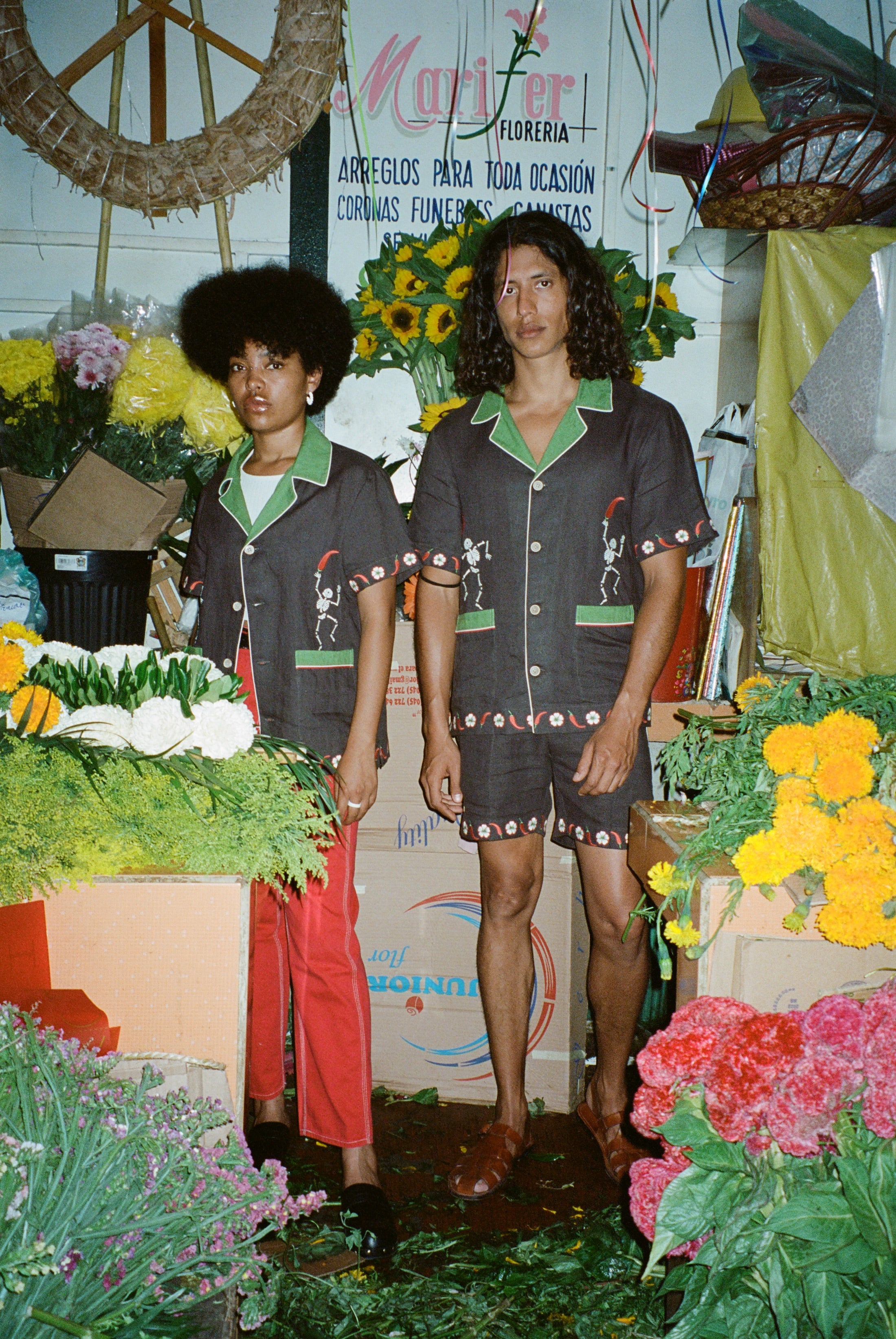 Man and woman standing in flower shop, each wearing brown picante shirt. The woman styles it with red pants. The man is wearing the matching shorts.