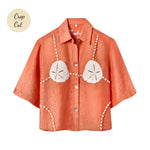 Orange short-sleeve button-up shirt with a relaxed fit. The shirt features an embroidered design resembling a sand dollar bikini top, with two large cream-colored sand dollars at the chest and shell bead strands extending from the shoulders, across the chest, and down the sides. Small sticker that reads "Crop Cut"