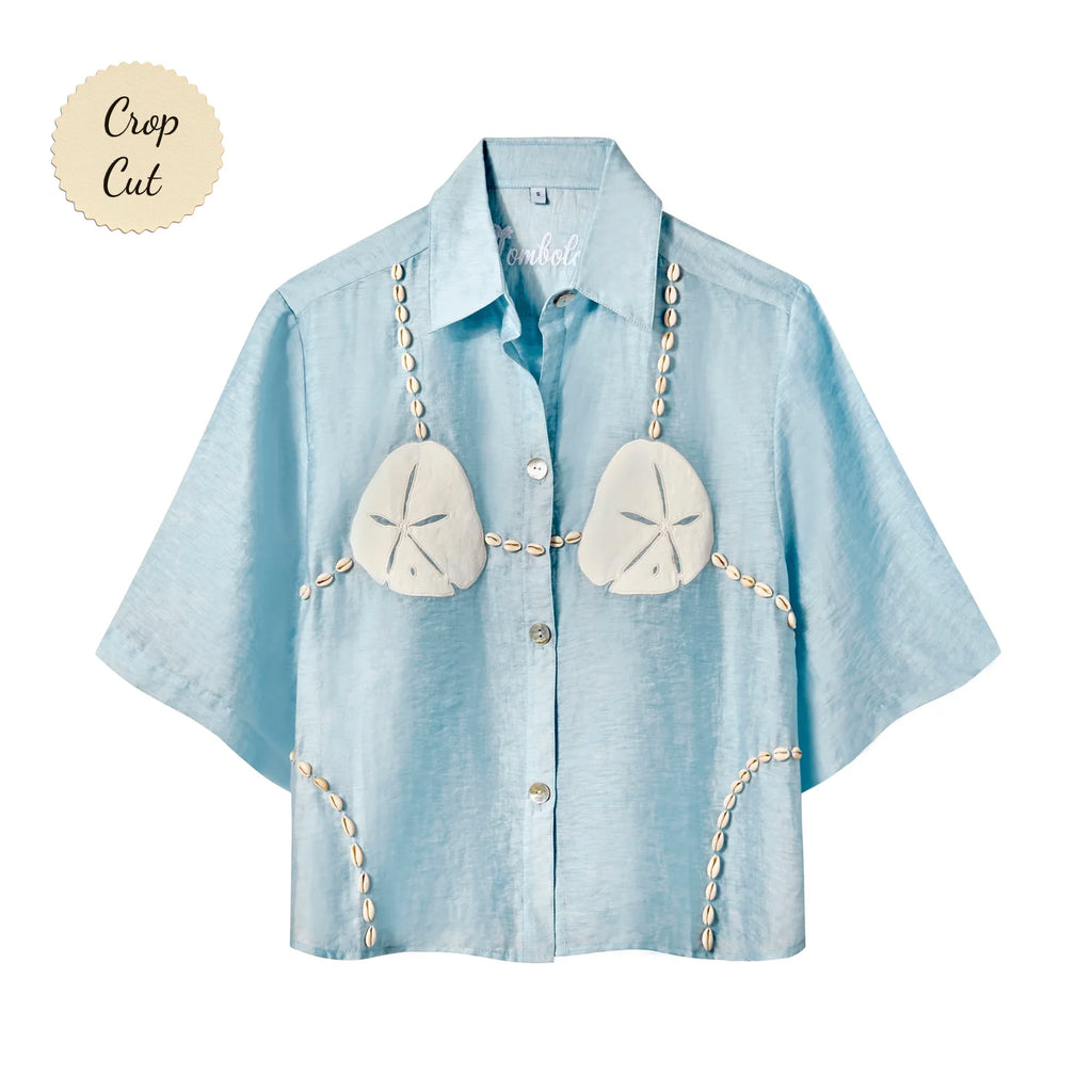 Light blue, short-sleeve button-up shirt with a relaxed fit. The shirt features an embroidered design resembling a sand dollar bikini top, with two large cream-colored sand dollars at the chest and shell bead strands extending from the shoulders, across the chest, and down the sides. Small sticker that reads "Crop Cut".