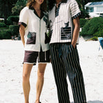 Male and female models on the beach with Female model on the left wearing the Tombolo 'Tombarolo' beige and purple linen set Male model on the right wearing Tombolo beige striped pop over shirt with matching striped pants 