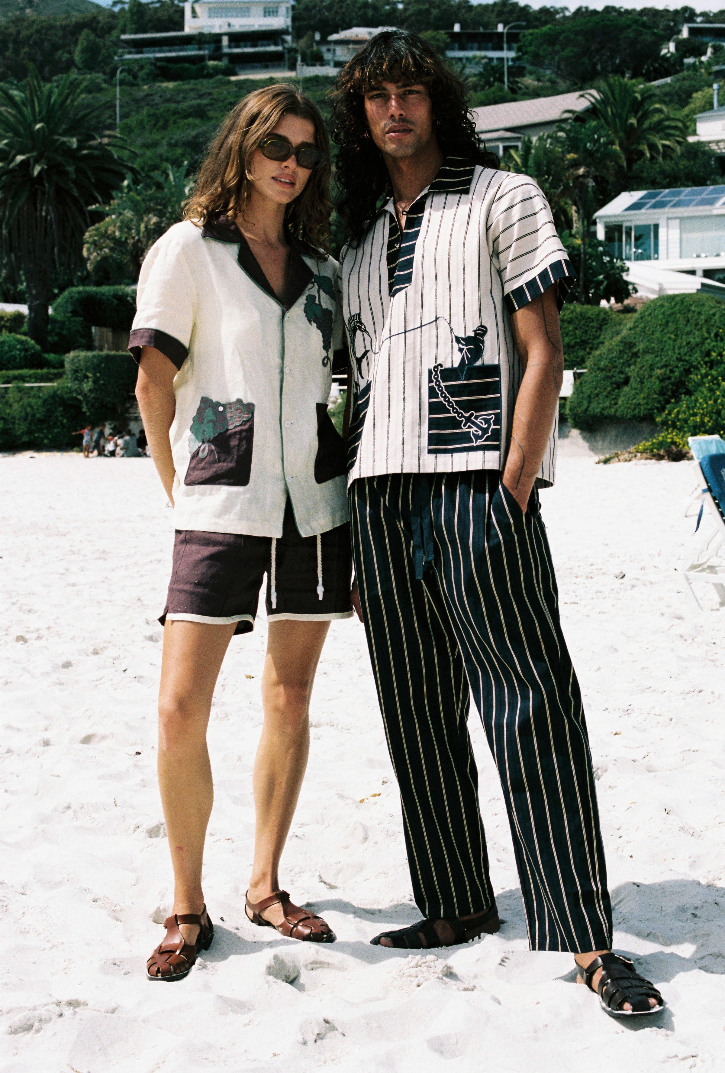Male and female models on the beach with Female model on the left wearing the Tombolo 'Tombarolo' beige and purple linen set Male model on the right wearing Tombolo beige striped pop over shirt with matching striped pants 