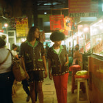 Man and woman walking in a market, each wearing the brown 'Picante' cabana shirt. The woman styles it with red pants. The man is wearing the matching shorts. 