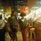 Man and woman walking in a market, each wearing the brown 'Picante' cabana shirt. The woman styles it with red pants. The man is wearing the matching shorts. 