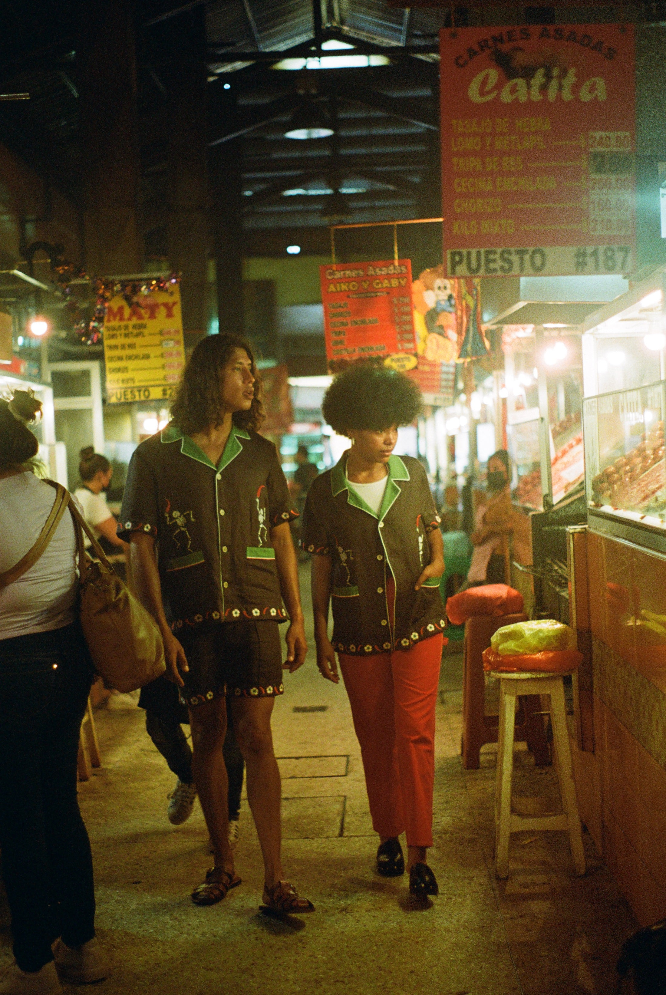 Man and woman walking in a market, each wearing the brown 'Picante' cabana shirt. The woman styles it with red pants. The man is wearing the matching shorts. 