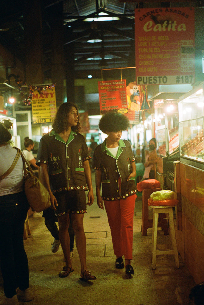 Man and woman walking in a market, each wearing the brown 'Picante' cabana shirt. The woman styles it with red pants. The man is wearing the matching shorts. 