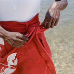 close up of woman at the beach wearing a white one piece bathing suit and tying the bow on her red skirt
