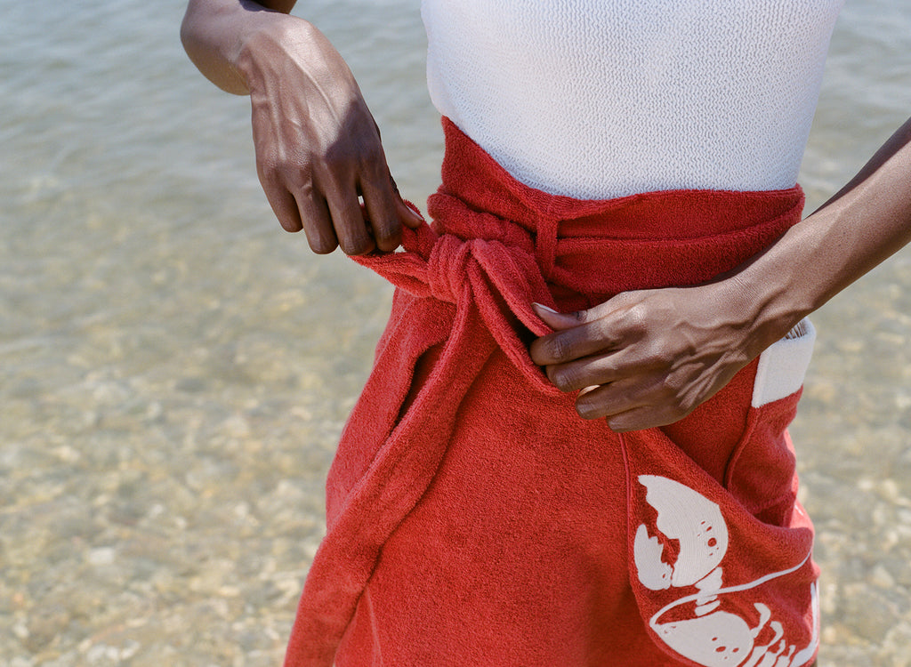 close up of woman at the beach wearing a white one piece bathing suit and tying the bow on her red skirt
