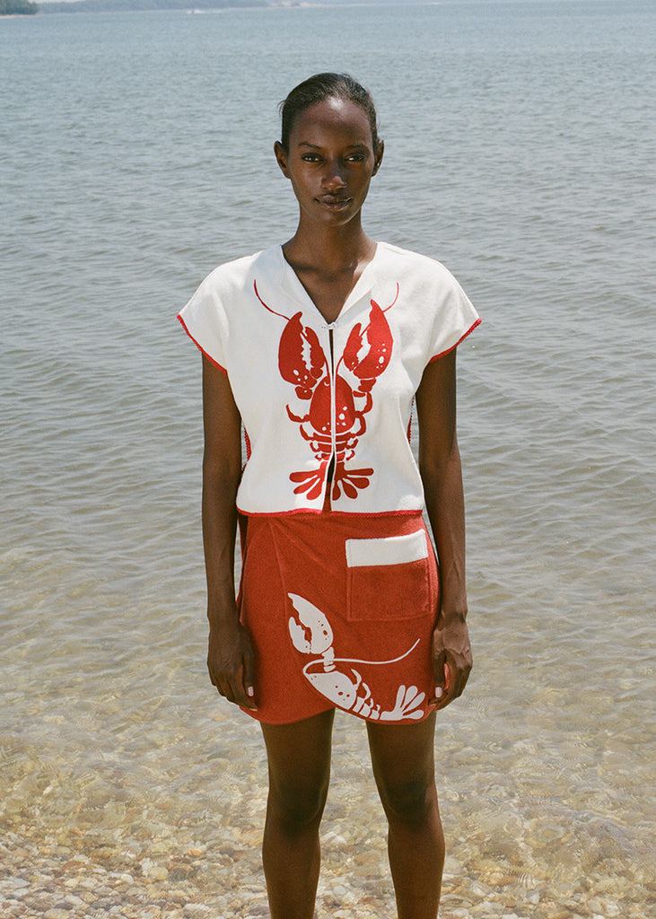woman at beach wearing white lobster bib shirt and red skirt, looking straight at the camera 
