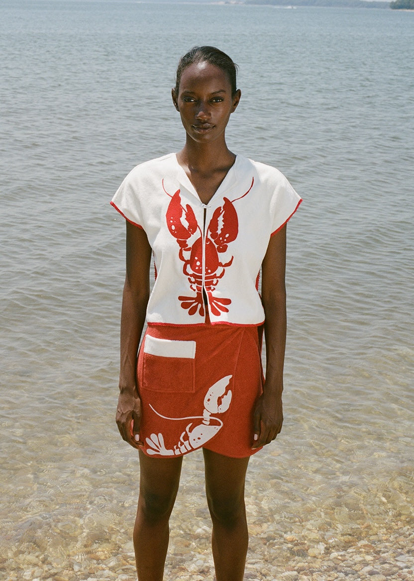 woman at beach wearing white lobster bib shirt and red skirt, looking straight at the camera 