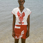 woman at beach wearing white lobster bib shirt and red skirt, looking straight at the camera 