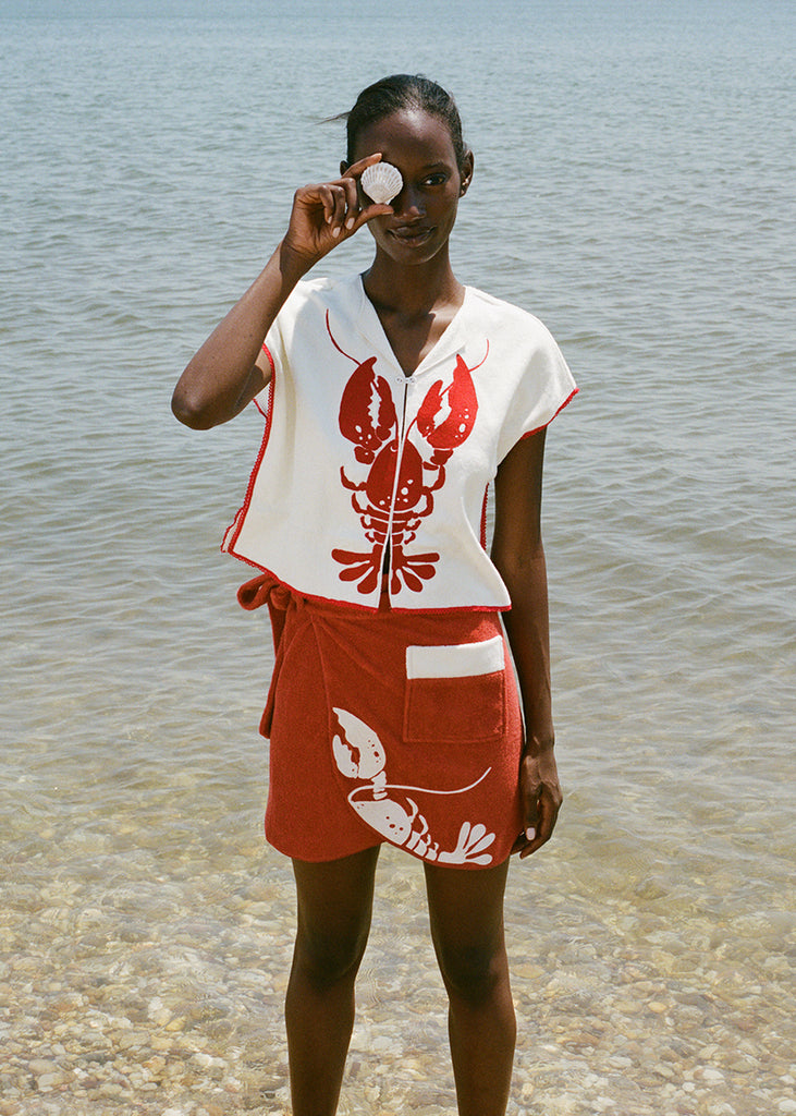 woman at beach wearing white lobster bib shirt and red skirt, smiling and holding a shell up to her eye 