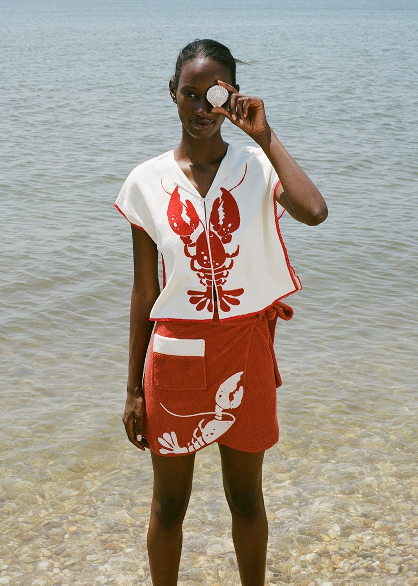 woman at beach wearing white lobster bib shirt and red skirt, smiling and holding a shell up to her eye 