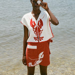 woman at beach wearing white lobster bib shirt and red skirt, smiling and holding a shell up to her eye 
