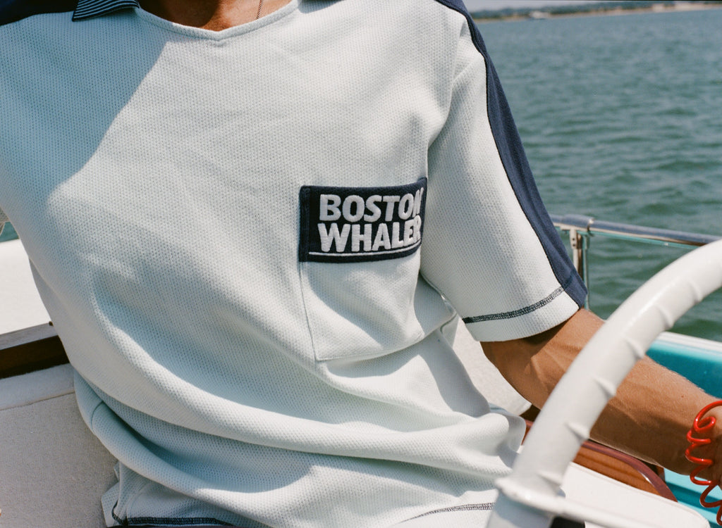 Close-up of a light blue cotton mesh shirt with navy sleeve panels and navy contrast stitching. The shirt features a navy chest pocket with white 'Boston Whaler' embroidery.