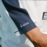 Close-up of a cotton mesh shirt with navy sleeve panels and navy contrast stitching. The sleeve features a small white 'Tombolo' logo, and part of the navy chest pocket with 'Boston Whaler' embroidery is visible.