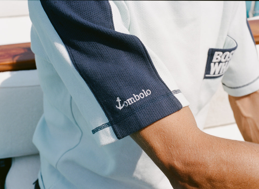 Close-up of a cotton mesh shirt with navy sleeve panels and navy contrast stitching. The sleeve features a small white 'Tombolo' logo, and part of the navy chest pocket with 'Boston Whaler' embroidery is visible.