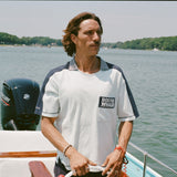 Man standing at the helm of a boat wearing a light blue cotton mesh shirt with navy shoulder panels and a striped ribbed collar. The shirt features a navy chest pocket with white 'Boston Whaler' embroidery and navy contrast stitching on the sleeves.
