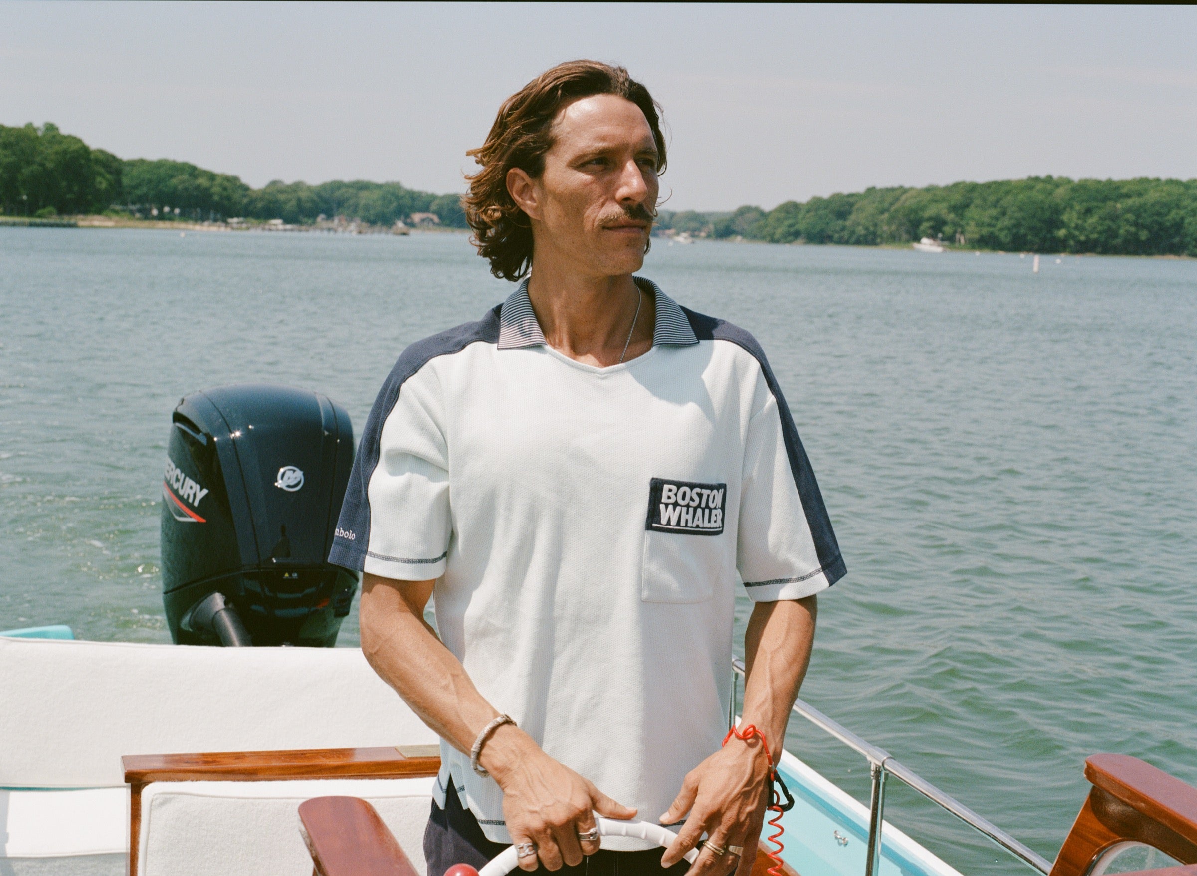 Man standing at the helm of a boat wearing a light blue cotton mesh shirt with navy shoulder panels and a striped ribbed collar. The shirt features a navy chest pocket with white 'Boston Whaler' embroidery and navy contrast stitching on the sleeves.
