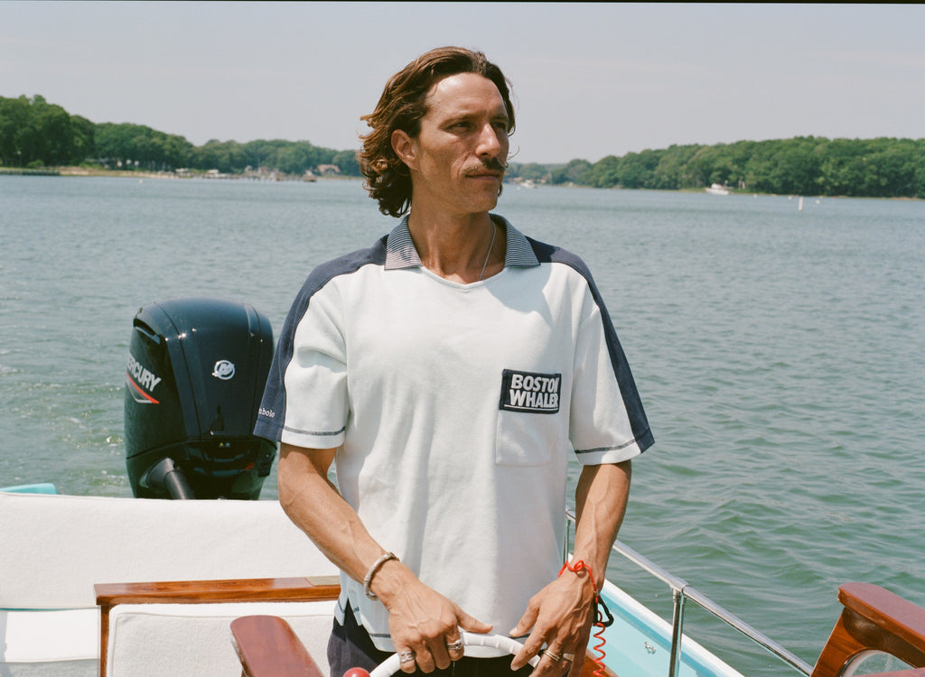Man standing at the helm of a boat wearing a light blue cotton mesh shirt with navy shoulder panels and a striped ribbed collar. The shirt features a navy chest pocket with white 'Boston Whaler' embroidery and navy contrast stitching on the sleeves.