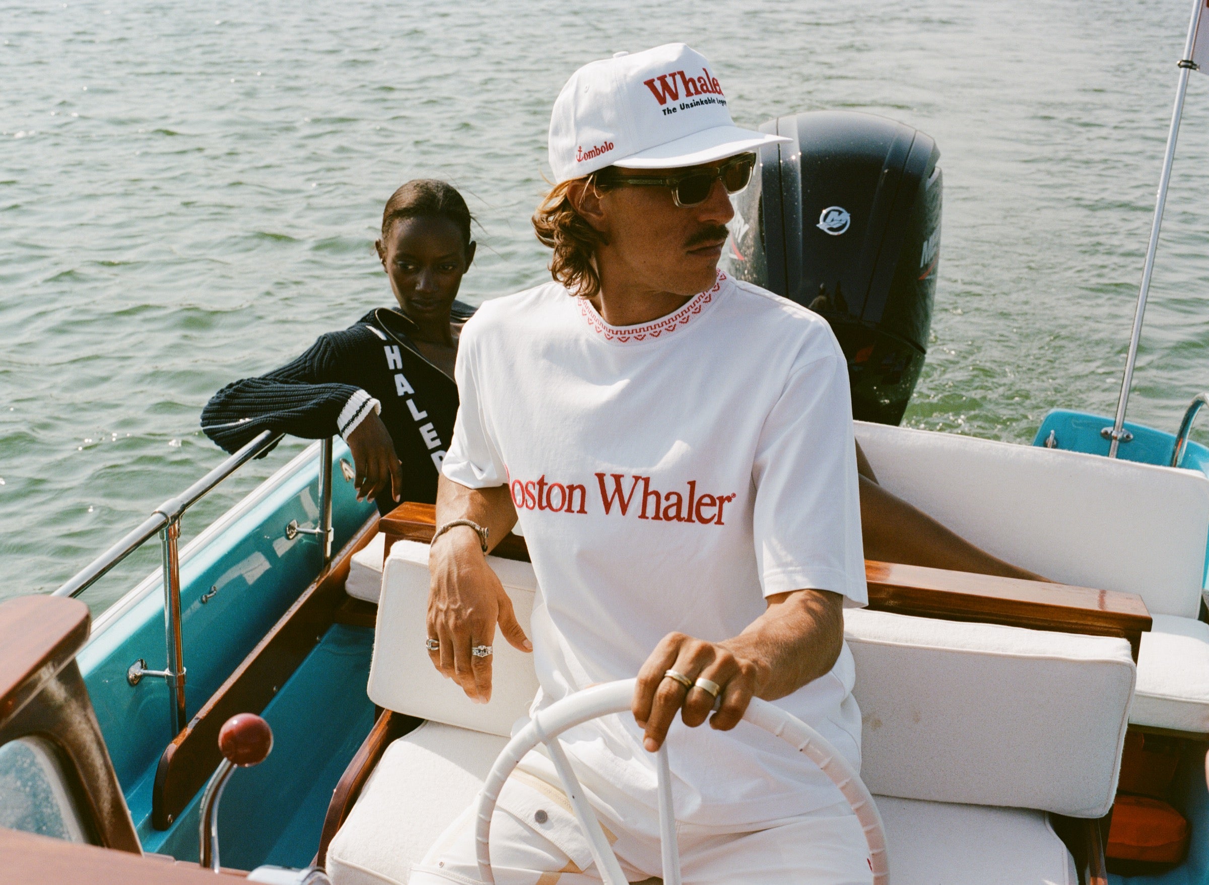 Man in a white five-panel cap and matching white shirt that reads 'Boston Whaler' steers a boat. The cap has red text reading 'Whaler' and smaller black text below reading 'The Unsinkable Legend.' A small red 'Tombolo' logo is on the side of the hat. A woman sits behind him wearing a navy sweater with white trim and bold white vertical lettering that reads 'WHALER.' 
