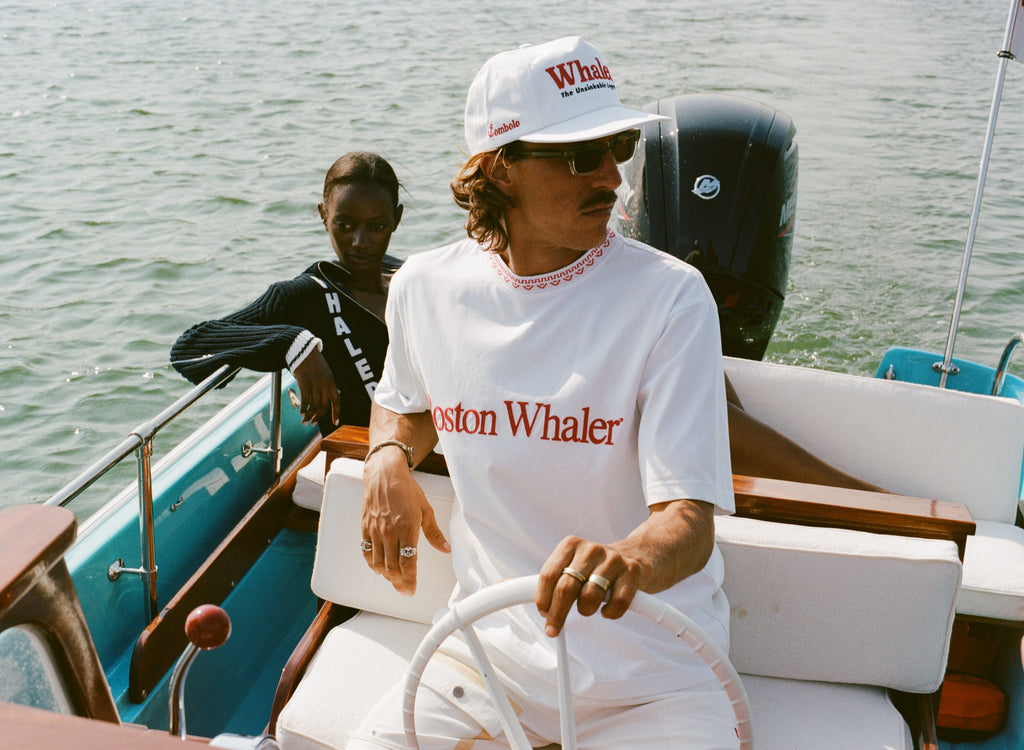 Man in a white five-panel cap and matching white shirt that reads 'Boston Whaler' steers a boat. The cap has red text reading 'Whaler' and smaller black text below reading 'The Unsinkable Legend.' A small red 'Tombolo' logo is on the side of the hat. A woman sits behind him wearing a navy sweater with white trim and bold white vertical lettering that reads 'WHALER.' 