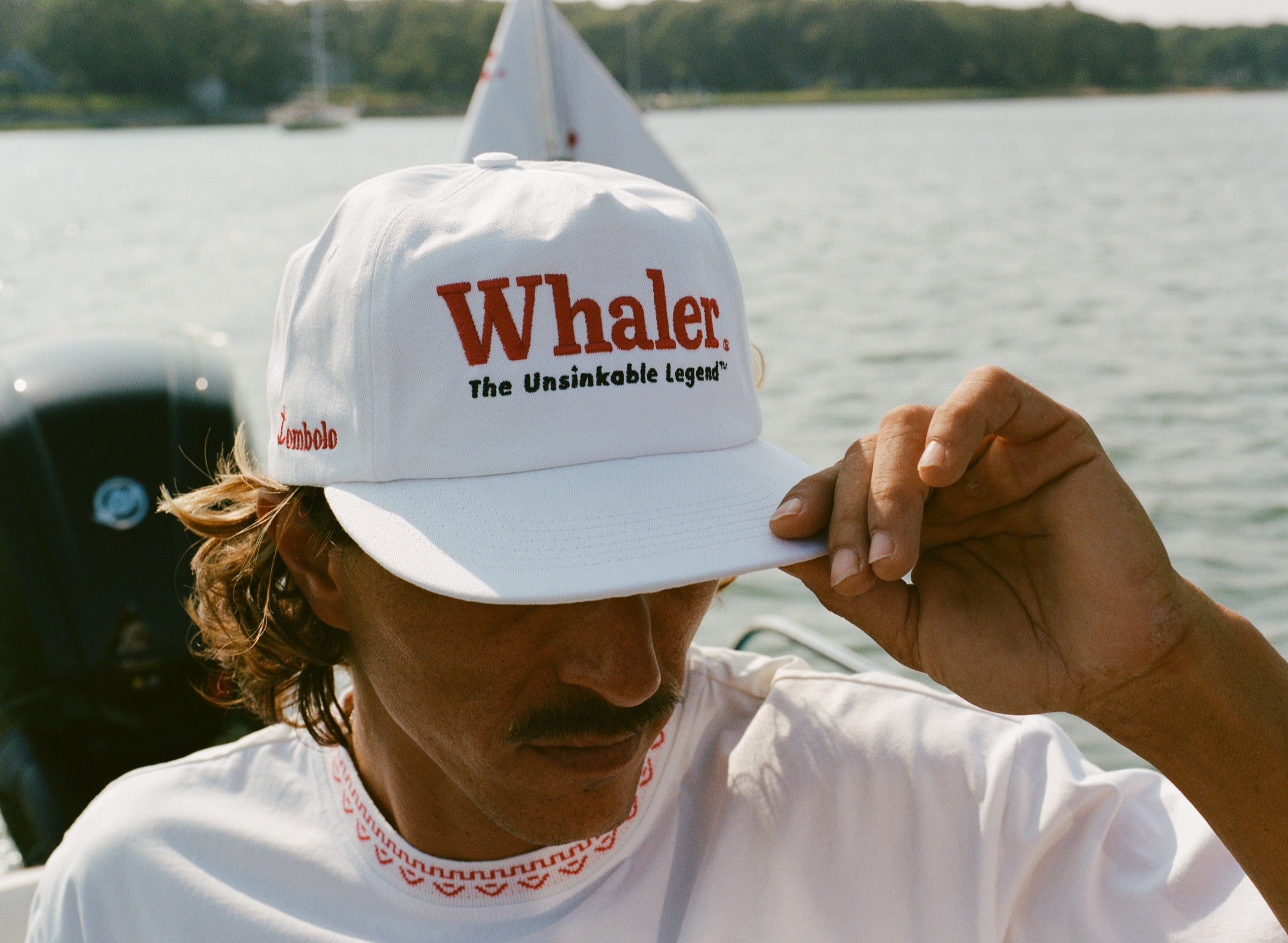 Close-up of a man holding the brim of a white five-panel cap with red text reading 'Whaler' and black text below reading 'The Unsinkable Legend.' A small red 'Tombolo' logo is embroidered on the side of the hat. He wears a white shirt with red decorative stitching along the collar.