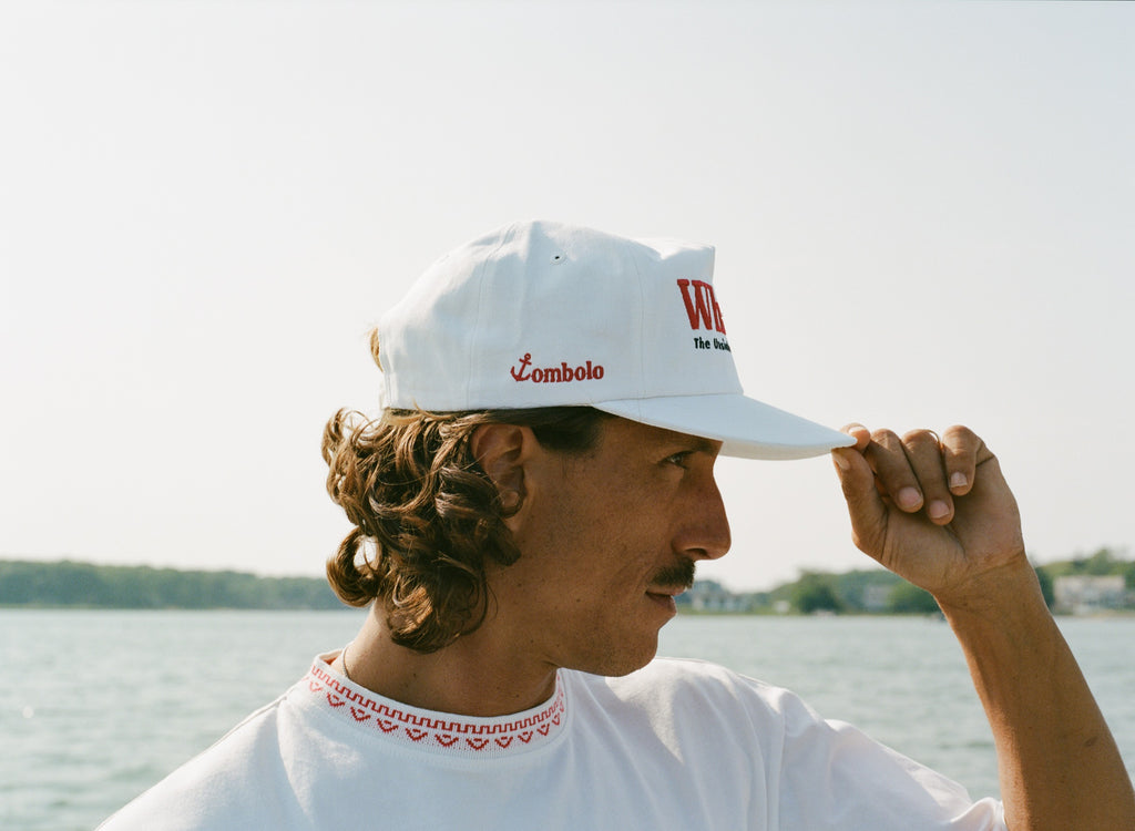Close-up of man on a boat wearing a white 'Whaler' cap with red and black embroidery and a white T-shirt with 'Boston Whaler' in red text. The side of the cap reads 'Tombolo' in red. 