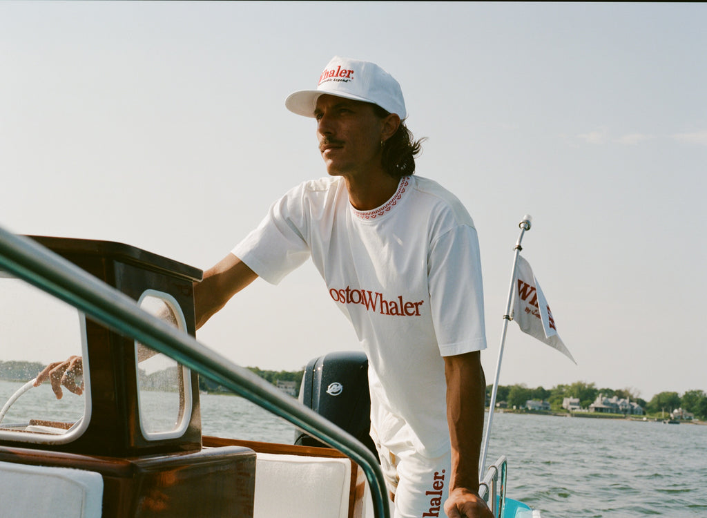 Man steering a boat while wearing a white five-panel cap with red text reading 'Whaler' and a matching white outfit with red 'Boston Whaler' branding. A white flag with the same logo is flying at the back of the boat.