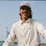 Man smiling while steering a boat, wearing a cream hooded jacket with tan trim and a white cap reading 'Whaler Unsinkable Legend.' A small Whaler flag waves in the background.