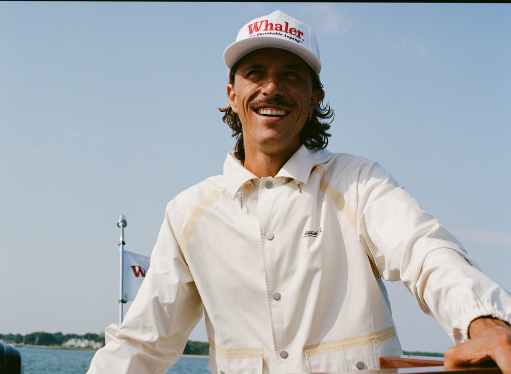 Man smiling while steering a boat, wearing a cream hooded jacket with tan trim and a white cap reading 'Whaler Unsinkable Legend.' A small Whaler flag waves in the background.