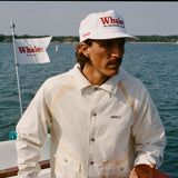 Man steering a boat in a cream jacket with tan trim and a white cap reading 'Whaler The Unsinkable Legend.' A Whaler flag waves behind him.
