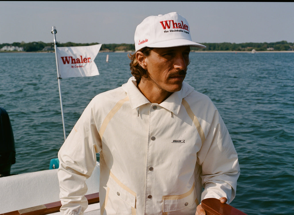 Man steering a boat in a cream jacket with tan trim and a white cap reading 'Whaler The Unsinkable Legend.' A Whaler flag waves behind him.