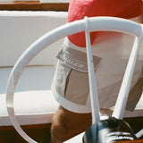 Close-up of a man wearing two-tone beige and off-white shorts with a flap pocket on the left featuring white 'Boston Whaler' embroidery. A red shirt is partially visible above the waistband. The shot is taken from behind a white boat steering wheel.
