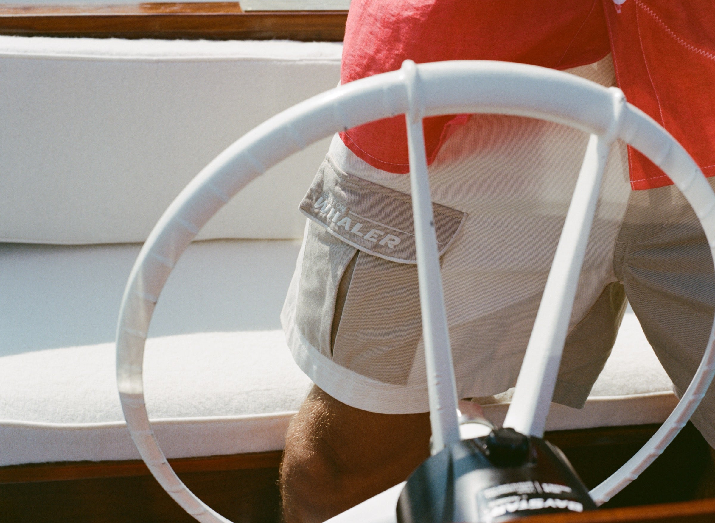 Close-up of a man wearing two-tone beige and off-white shorts with a flap pocket on the left featuring white 'Boston Whaler' embroidery. A red shirt is partially visible above the waistband. The shot is taken from behind a white boat steering wheel.