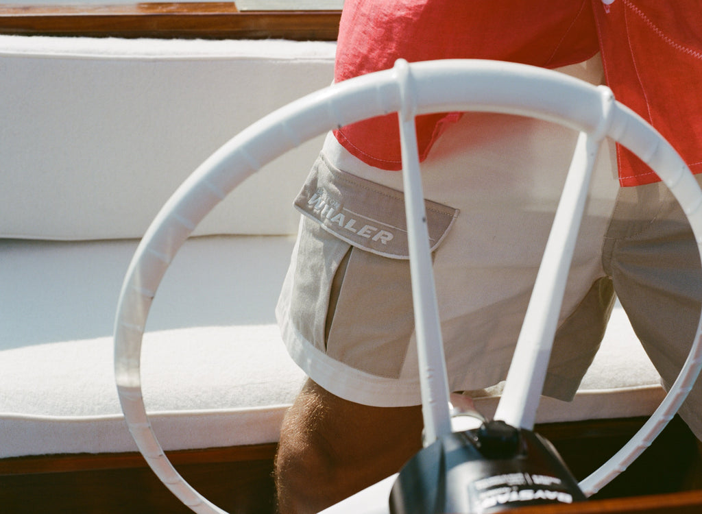 Close-up of a man wearing two-tone beige and off-white shorts with a flap pocket on the left featuring white 'Boston Whaler' embroidery. A red shirt is partially visible above the waistband. The shot is taken from behind a white boat steering wheel.